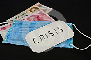 Inscription `crisis` on the sticker, medical mask and Chinese yuan on a black background.