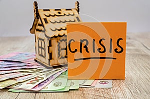 The inscription `crisis` on the sticker, Ukrainian hryvnia and a small wooden house. The concept of crisis, rising or falling real