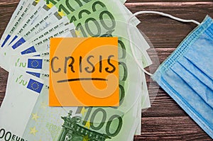 The inscription `crisis` on a yellow sticker with euro banknotes and a medical mask. Crisis concept. View from above.