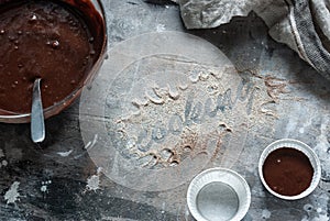 The inscription flour `Cooking` on a gray wooden messy background. Chocolate muffin dough. Cozy culinary cover, concept, cooking