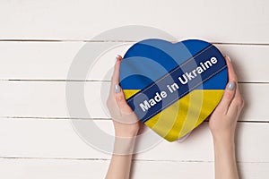 Inscription Made in Ukraine, the flag of Ukraine. Female hands holding a heart shaped box. White wooden table on a background.