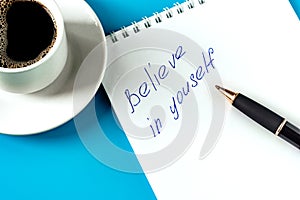 the inscription on the white sheet in the notebook believe in yourself. Black ballpoint pen on white notepad and a cup of coffee