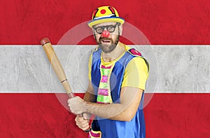 An insecure clown holds a bat in his hands against the background of the flag of Austria