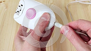 Insert wire into device. White epilator. Female hands turn on electric gadget to remove hair. Close-up, wooden background