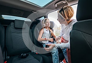 Inside the car photo of a mother fastening with safety auto belt her little daughter girl sitting in child seat. Girl listening