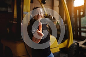 Inside the warehouse, a young woman stands in front of a yellow forklift and shows her thumbs up. Finger accent, out of focus