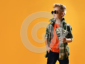 Insolent daring rich teenager boy in round sunglasses, checkered plaid shirt and jeans stands holding bundle of cash