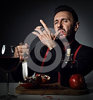 Insolent man in black shirt and suspenders at table with fresh pomegranate and juice, holds knife and licks his finger