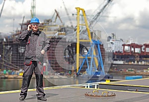 Inspector in coveralls and hardhat standing with mobile phone on harbor wharf.