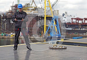 Inspector in coveralls and hardhat standing with tablet on harbor wharf.