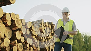 An inspector in a white helmet examines a pile of felled trees. Passes about felled trees. Checks the condition and size