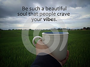 Inspirational motivational quote - Be such a beautiful soul that people crave your vibes. With person holding coffee cup in field.