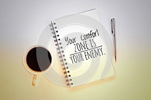 Inspirational motivational quote on notebook - Your comfort zone is your enemy. With cup of morning coffee, book and pen on table.