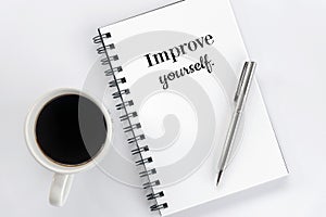 Inspirational motivational words - Improve yourself, on a spiral notepaper book with silver pen and a cup of black morning coffee.