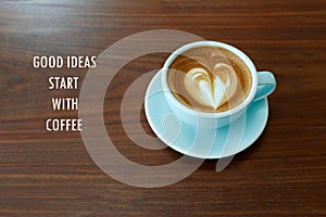 Inspirational positive quote `Good ideas start with coffee` with heart shape latte coffee and wooden desk background.