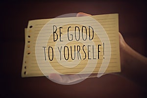 Inspirational quote - Be good to yourself. With a paper note in young woman hand on light brown background. Reminder, note to self