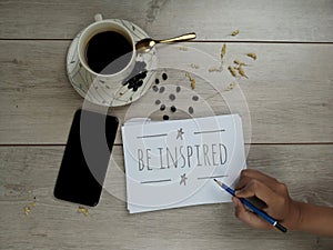 Inspirational quote - Be inspired. With a cup of white coffee,  phone and note on spiral white notebook on wooden table background