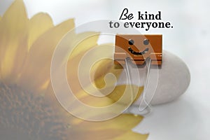 Inspirational quote - Be kind to everyone. With smile symbol on yellow paper clip and blurry sunflower on white background.