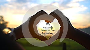 Inspirational quote - We can only learn to love by loving. On person making heart shape for love sign against sunset.