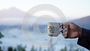 Inspirational quote - Do more of what makes your soul happy. With person holding cup of coffee on foggy mountain background.