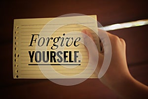 Inspirational quote - Forgive yourself. With a paper note in young woman hand on light brown background. Reminder and note to self