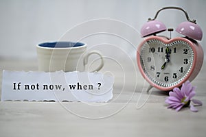 Inspirational quote - If not now, when. With a cup of coffee, pink clock alarm table and purple daisy flower on white wooden table photo