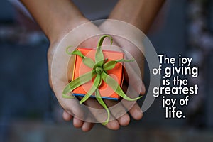 Inspirational quote - The joy of giving is the greatest joy of life. With a small cute gift box in young woman hands.