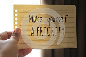 Inspirational quote - Make yourself a priority. With a paper note reminder in young woman hand against back light background.