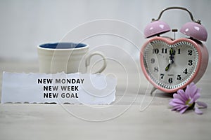 Inspirational quote - New Monday, New Week, New Goal. With note a cup of coffee, pink desk clock and purple daisy flower.