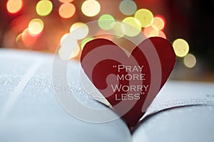 Inspirational quote on a red heart - Pray more, worry less. On open bible book page and colorful bokeh lights background.