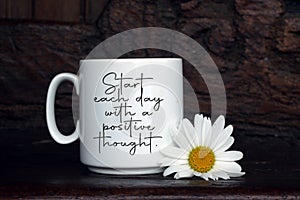 Inspirational quote - Start each day with a positive thought. With white cup of coffee or tea and daisy flower on the table.