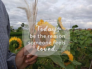 Inspirational quote - Today, be the reason someone feels loved. With blurry background of sunflowers garden, hand holding flower.v