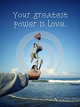 Inspirational quote - Your greatest power is love. With person holding dry red rose in hand on blue summer sky background.