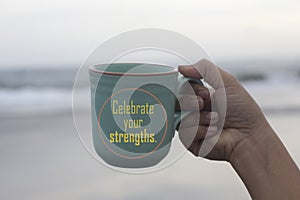 Inspirational text message on a coffee cup - Celebrate your strengths. With person holding a cup of coffee in hand on white beach.