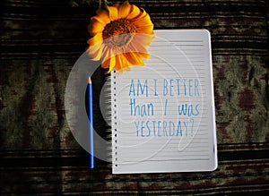 Inspiratonal motivational quote - Am i better than i was yesterday.  Notes on white paper notebook with blue pen, sunflower