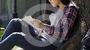 Inspired female student of arts faculty drawing, sitting under tree on campus