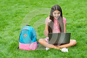 Inspired happy child using computer for online school study sitting in park on green grass with backpack, education