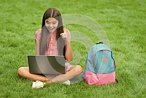 inspired happy child using computer for online school study sitting in park on green grass with backpack, education