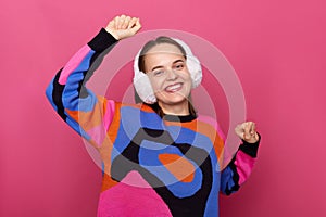 Inspired jocund girl wearing colorful jumper and fur earmuffs, dancing isolated on pink background, gorgeous young female model