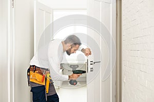 Installation of a lock on the front wooden entrance door. Portrait of young locksmith workman in blue uniform installing