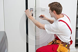 Installation of a lock on the front wooden entrance door. Portrait of young locksmith workman in blue uniform installing