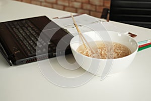 Instant noodles in a bowl near laptop on white working desk in office