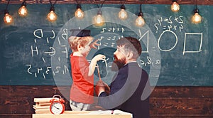 Instructive conversation concept. Child in graduate cap listening teacher, chalkboard on background, rear view. Teacher