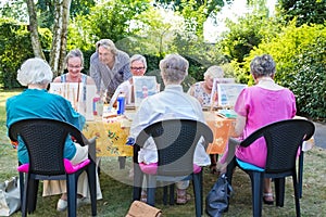 Instructor helping a group of senior retired ladies at art class seated around a table painting outdoors in a garden or park.