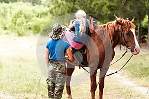 The instructor helps the girl to saddle a brown horse in the forest. Children`s equestrian camp. Summer sports camp for children.