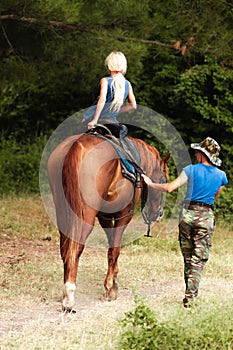 The instructor helps the girl to saddle a brown horse in the forest. Children`s equestrian camp. Summer sports camp for children.