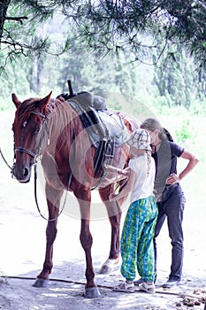 The instructor helps the girl to saddle a brown horse in the forest. Children`s equestrian camp. Summer sports camp for children.