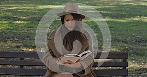 Intelligent Caucasian female student in brown hat and coat sitting on the bench and reading book. Beautiful European