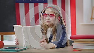 Intelligent relaxed schoolgirl in headphones reading book smiling sitting in school classroom at desk. Portrait of