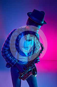 Intense musician, African-American man in hat playing saxophone in vivid lighting against pink-blue background.